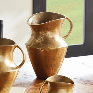 The ELAINA DECO PITCHER by NAPA HOME AND GARDEN features a golden, textured cast aluminum design with a large, curved handle and a slightly flared rim. Its surface boasts a matte, hammered finish that offers a rustic yet elegant look, ideal for adding charm to any transitional space. The decorative pitcher is showcased against a plain white background.