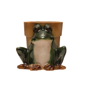 The Creative Coop Frog Planter, featuring a shiny green finish and a detailed texture with glossy eyes, sits adorably in front of a plain brown pot. This quirky vessel, designed for small plants, boasts a charming reactive glaze finish.