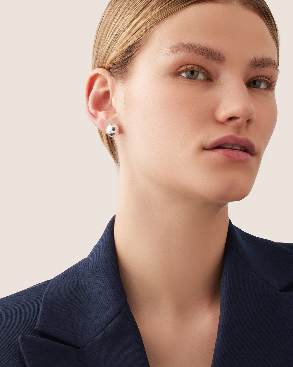 A person with short blonde hair is wearing a dark blazer and the JENNY BIRD Aurora Stud in Silver earrings. They have a neutral expression, looking slightly to the side against a plain, light background.