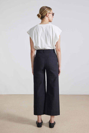 In a minimalist setting, an individual confidently poses against a neutral background, wearing sleek sunglasses, a white T-shirt, the APIECE APART - CLASSIC MERIDA PANT in black high-waisted wide-legged style, and stylish black loafers.