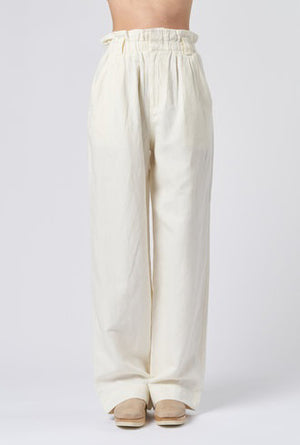 The individual is wearing the SON VIDA PANT by APIECE APART. These high-waisted pants are loose-fitting and crafted from organic cotton, featuring pockets and stitching details on the back. Designed for comfort, they include an elastic waistband and are showcased in a rear-view photograph against a plain backdrop.