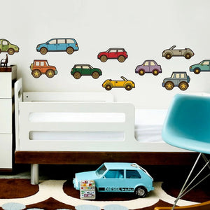 A child's room featuring a white crib-like bed, blue chair, and carpet showcasing a brown, white, and orange circular pattern. SIMPLE SHAPES - CAR REMOVEABLE WALL DECALS by SIMPLE SHAPES in various colors are adhered to the wall above the bed. A blue toy car is positioned on the floor near the bed.