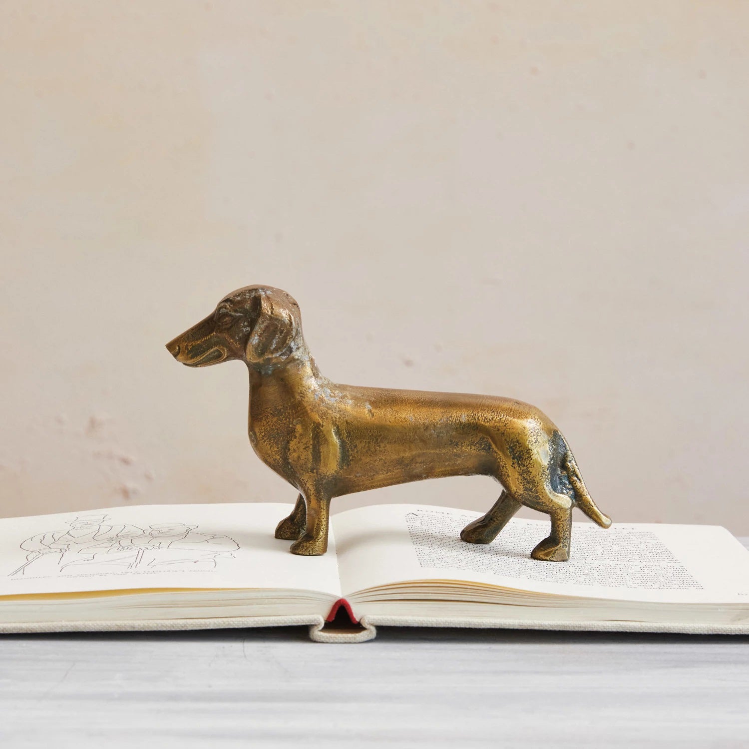 A polished CAST ALUMINUM DACHSUND by CREATIVE COOP stands atop an open book, making the perfect dachshund accent for dog lovers decor. The book lies on a light-colored surface, with text and illustrations visible on the pages. The background is blurred to focus attention on the vintage brass-like finish of the figurine and the book.