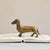 A polished CAST ALUMINUM DACHSUND by CREATIVE COOP stands atop an open book, making the perfect dachshund accent for dog lovers decor. The book lies on a light-colored surface, with text and illustrations visible on the pages. The background is blurred to focus attention on the vintage brass-like finish of the figurine and the book.