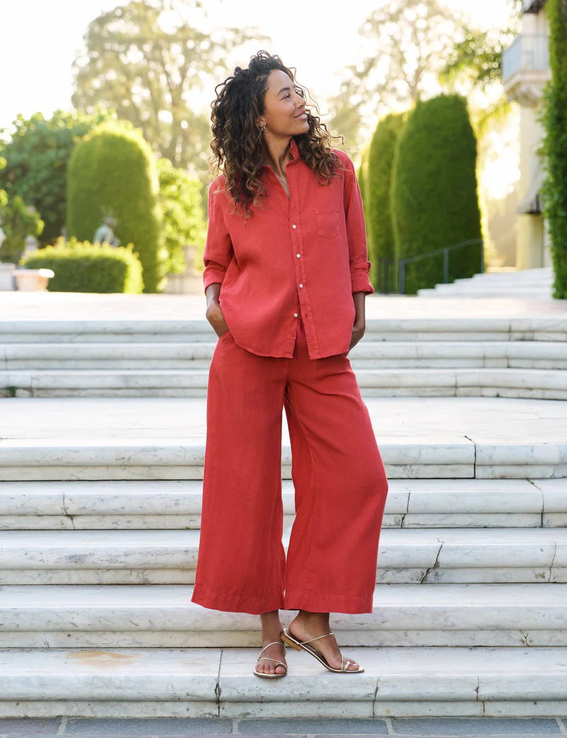On marble steps outdoors, a woman with curly hair wears gold sandals and the FRANK &amp; EILEEN Maisie Wide Leg Pull On Pant from the Tuscany Getaway Set in Summer Red. She stands with hands in her pockets, framed by green trees and manicured bushes.