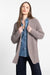 A woman with long, straight hair is wearing the KINROSS CASHMERE - LUXE GARTER STITCH CARDIGAN in a chunky beige knit made of 100% cashmere, layered over a blue denim shirt and paired with dark jeans. She stands against a plain white background, holding the sides of the cardigan with a relaxed expression.