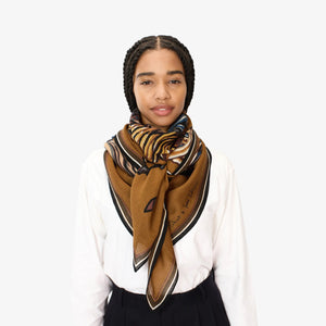 A person with braided hair is wearing a white shirt and the INOUI EDITIONS WESTERN SQUARE SCARF by INOUI, a patterned brown wool accessory. The plain, light-colored background accentuates the unisex appeal of their outfit.