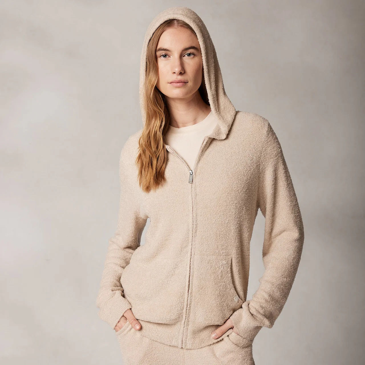 A person with long hair, wearing Kashwere's KASHWERE - RELAXED HOODIE made from Kash-ease yarn, has their hands in the front pockets of a light beige zip-up. The hood is up against a neutral, textured background. This wardrobe staple from KASHWERE looks both comfortable and casual.