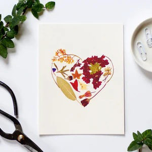 The CARD HEART from PETAL PEOPLE PRESS is a hand-crafted Valentine's Day card adorned with a heart made from pressed flowers and leaves. It is elegantly displayed on a white surface next to an envelope, a small bunch of red flowers, and a single green leaf. A floral stamp adds the perfect finishing touch, making it an ideal botanical art card for any occasion.