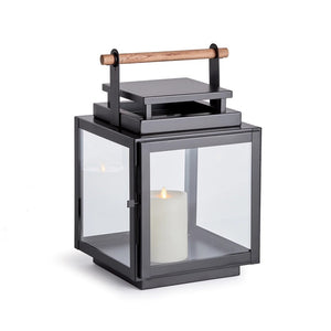 The ADWIN OUTDOOR LANTERN by NAPA HOME AND GARDEN is a modern black metal outdoor lantern with a teak handle and wide glass panes. Inside, it holds a large white pillar candle that is lit, casting a soft, warm glow. The lantern features a sleek, minimalist design and sits on a flat surface.