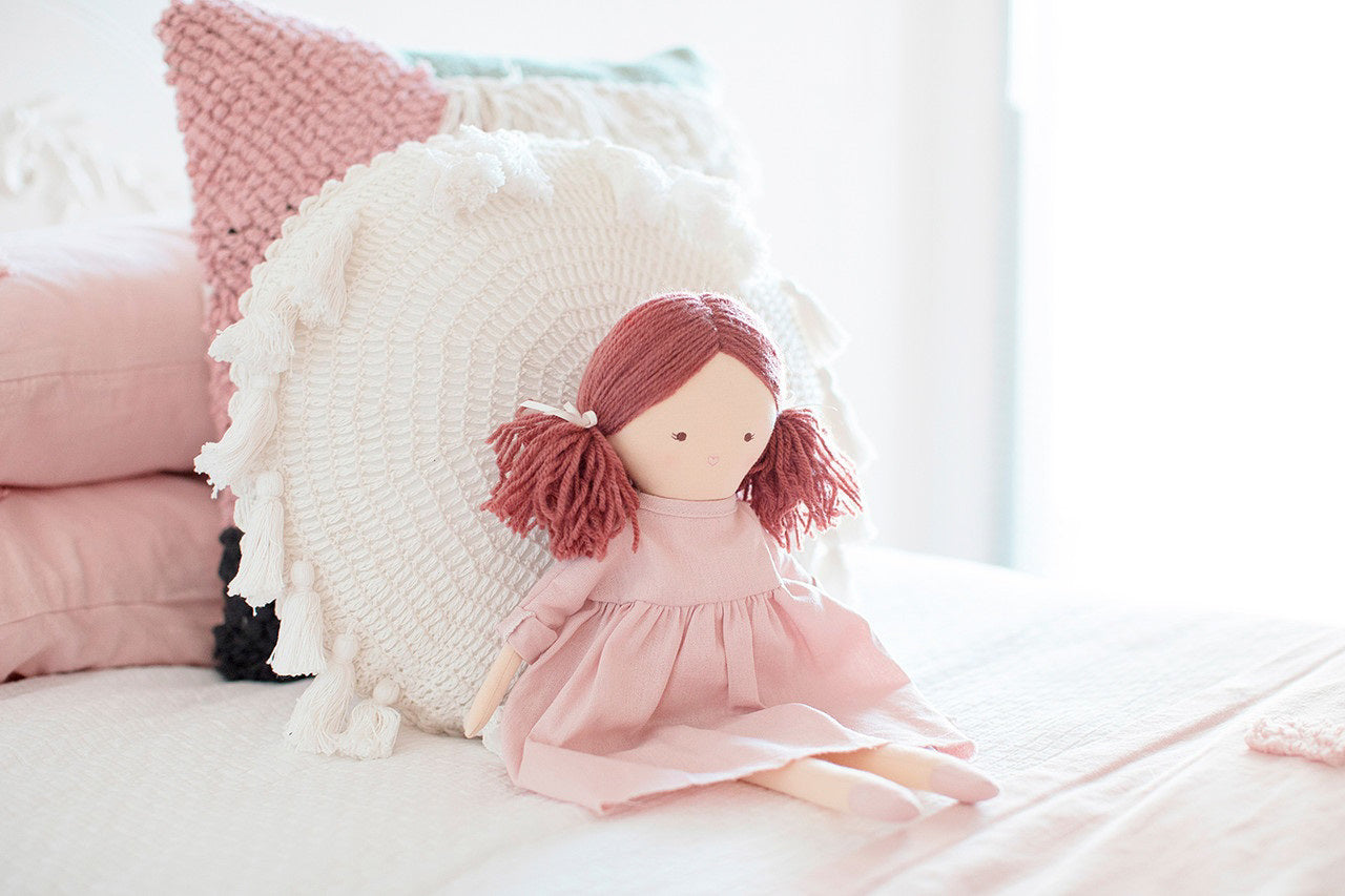 The ALIMROSE - MATILDA DOLL PINK by ALIMROSE is a soft fabric doll with dark red yarn hair styled in pigtails and an embroidered face. She is dressed in a light pink linen dress with long sleeves and matching pink shoes. The removable dress allows for easy outfit changes, adding to the fun.