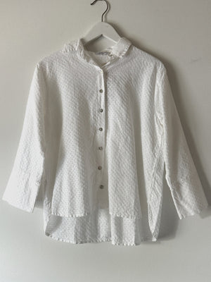 The CUT LOOSE - EASY CROP SHIRT, a white, long-sleeved blouse with a subtle dotted texture, is hanging on a white hanger against a plain wall. This elegant piece from CUT LOOSE features a classic collar, button-up front, and a hi-low hem for added style.