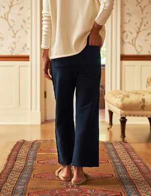 A person wearing a white long-sleeve shirt, FRANK & EILEEN's BELLA ITALIAN SWEATPANT IN BRITISH ROYAL NAVY, and slippers stands on a patterned rug in a cozy room with ornate wallpaper, wainscoting, and antique furniture. The person's back is facing the camera.