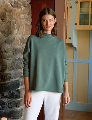 A person with shoulder-length brown hair is wearing the FRANK & EILEEN Effie Long Sleeve Funnel Neck Capelet in rosemary and white pants, smiling by a stone wall in a room with colorful cabinets, exuding the cozy vibe of a winter warmer.