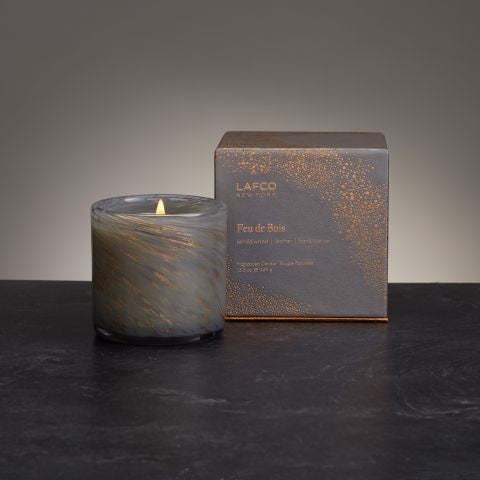 A LAFCO - Feu de Boise Holiday Candle, nestled in a marbled glass holder and infused with the essence of Virginia Cedarwood, rests on a dark surface beside its sophisticated gray and gold textured box inscribed with "LAFCO New York, Feu de Bois, woodfire." The setting is gently illuminated to emphasize the candle's warm glow.