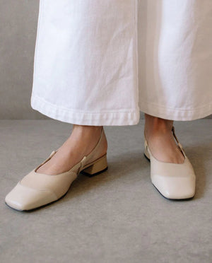 A person exudes workwear chic, wearing wide-leg white pants and beige NOAH SLIDE SANDALS by ALOHAS, standing confidently on a gray floor.