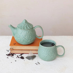 A 30 oz. embossed stoneware teapot with strainer from Creative Coop, featuring intricate leaf patterns, sits alongside a matching mug on a stack of books. Nearby, a tea bag and loose tea leaves are scattered on a white surface. The reactive glaze shimmers subtly against the simple beige wall in the background.