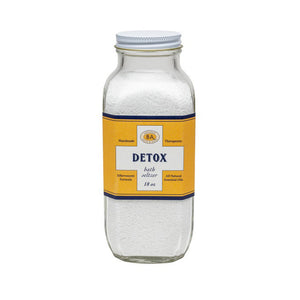 A clear glass jar with a white lid contains white bath salts. The jar's yellow and blue label reads: "BAUDELAIRE - DETOX BATH SELTZER 18 OZ." Additional text indicates it is handmade, therapeutic, all-natural with essential oils and natural ingredients, featuring a proprietary formula.