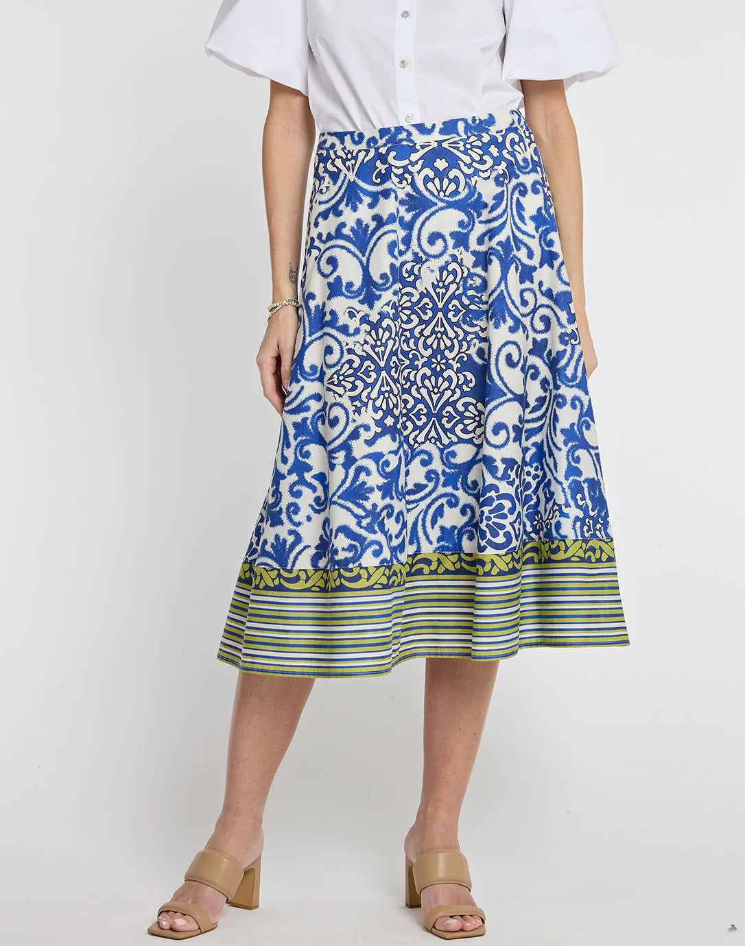 A person is modeling a white blouse with puffed sleeves and the HINSON WU - GLORIA SKIRT IN CERAMIC TILE PRINT, featuring a blue and white pattern with green and yellow striped accents at the hem. They are wearing beige open-toed, block-heeled sandals.