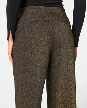 A person is wearing the SPANX AIRESSENTIALS WIDE LEG SHIMMER PANT in black along with a zip-up top. They stand casually with their hair tied back, sporting black and white sneakers, all set against a plain white background.