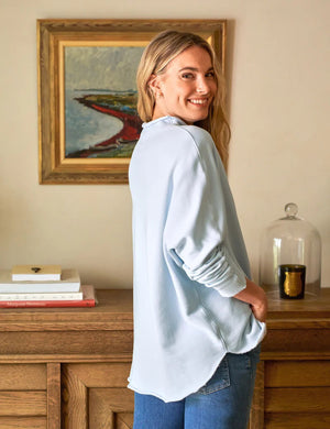 A woman with long hair, wearing a FRANK & EILEEN Effie Long Sleeve Funnel Neck Capelet in Ice and jeans, smiles over her shoulder. She stands in a room featuring a wooden sideboard, books, a candle, and a framed painting on the wall.