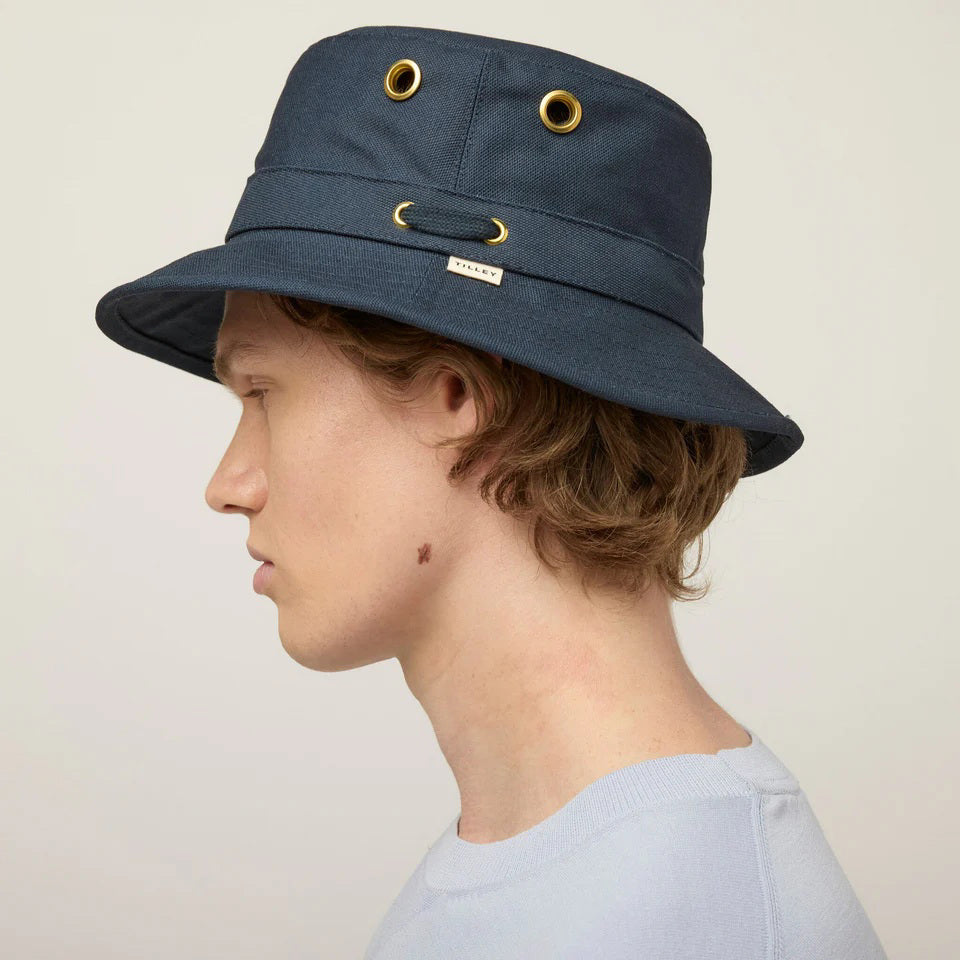 A young man faces forward against a plain background, wearing the TILLEY - ICONIC T1 BUCKET HAT in navy blue with gold side buttons and a light blue crew-neck shirt—a classic UPF 50+ sun protection style by TILLEY.