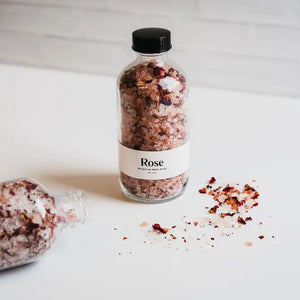 A transparent glass bottle containing a blend of pink and white salt crystals and rose petals, enriched with organic rose and lavender essences. The bottle features a white label that reads "BOTANICAL BATH SOAK - ROSE" from GIFTEN MARKET and is topped with a black cap. Set against a white background, it promises an indulgent bathing experience.