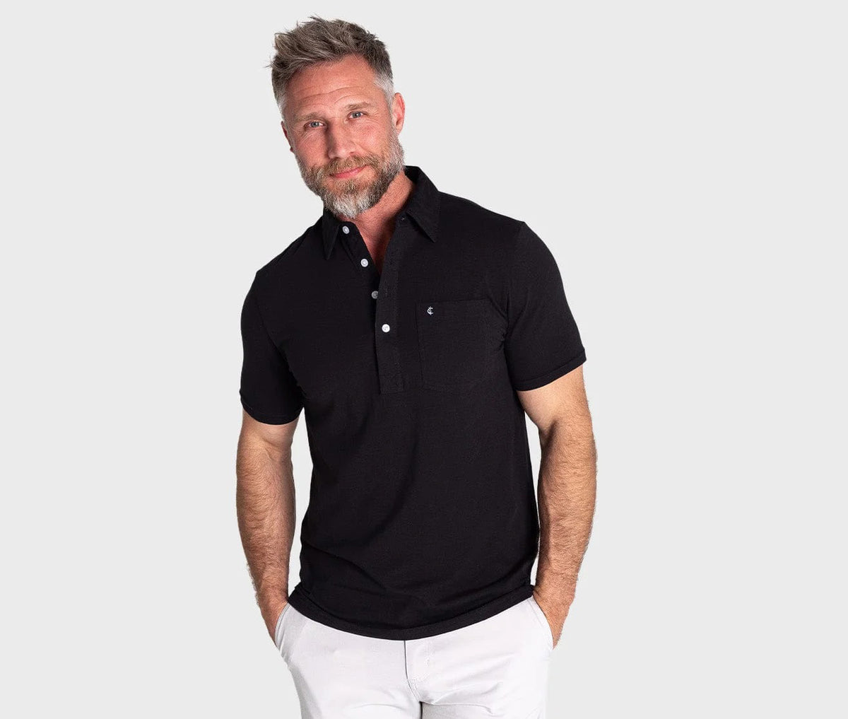 A man with short gray hair and a beard wears the CRIQUET - TOP SHELF PLAYERS SHIRT IN BLACK TOP by CRIQUET and white pants, standing with hands in his pockets against a plain light gray background.