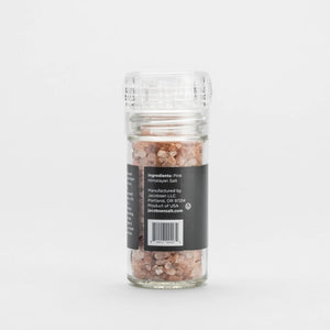 The JACOBSEN SALT CO. GRINDER PINK HIMALAYAN SALT, a clear glass grinder with a transparent plastic top, stands against a plain white background. The black label on this refillable grinder lists the ingredients and manufacturer details.