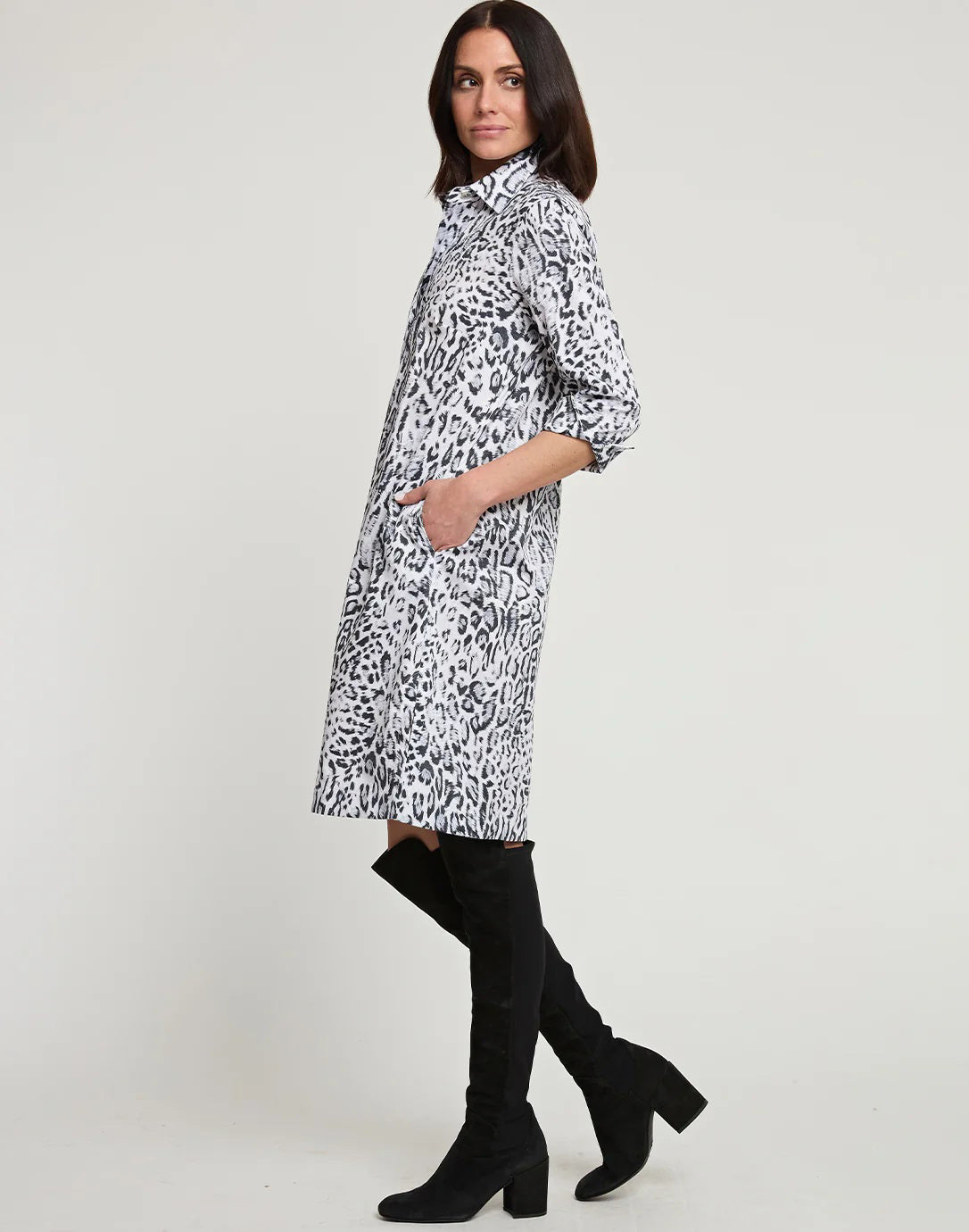 A woman with shoulder-length dark hair is wearing a knee-length HINSON WU - CHARLOTTE 3/4 Sleeve Snow Leopard Sateen Dress. She is also sporting black knee-high boots and has her hands in the dress pockets. The background is plain white.