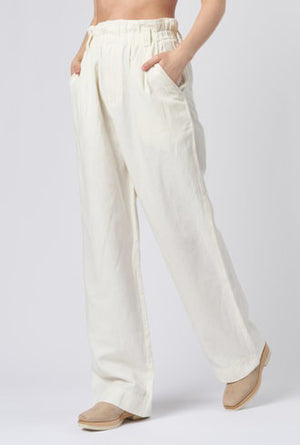 The individual is wearing the SON VIDA PANT by APIECE APART. These high-waisted pants are loose-fitting and crafted from organic cotton, featuring pockets and stitching details on the back. Designed for comfort, they include an elastic waistband and are showcased in a rear-view photograph against a plain backdrop.