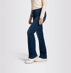 A person wearing a long-sleeve, light-colored ribbed top and MAC's DREAM BOOT AUTHENTIC DENIM boot cut jeans in dark blue with a slight flare at the bottom. They are also wearing white sneakers and have one hand in their pocket, standing against a plain, light gray background.