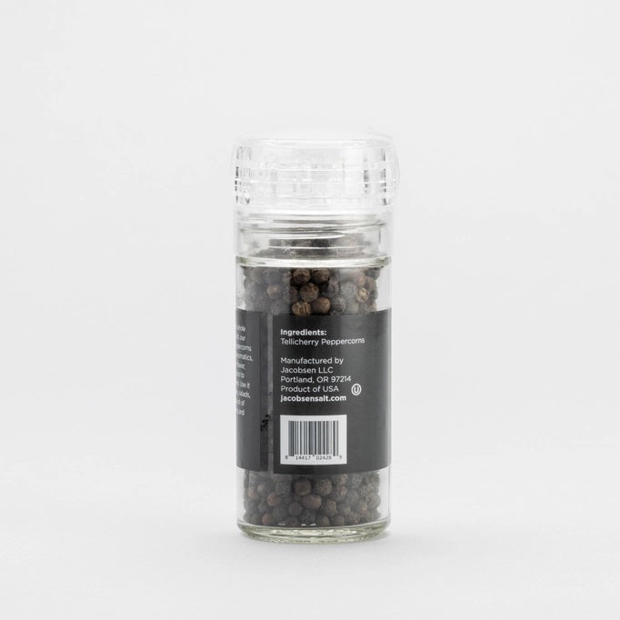 A JACOBSEN SALT CO. grinder, filled with Tellicherry peppercorns, sits gracefully on a light-colored surface against a plain, light gray background. The clear glass bottle bears a striking label in black with white and orange text.