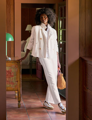 In a warmly lit hallway, a woman with curly hair wears the ultra-sophisticated FRANK & EILEEN - BELLA Italian Sweatpant in Vintage White, featuring an adjustable drawstring waist. She holds a brown bag over her shoulder and stands near elegantly designed wooden furniture in front of a glass-paned door.