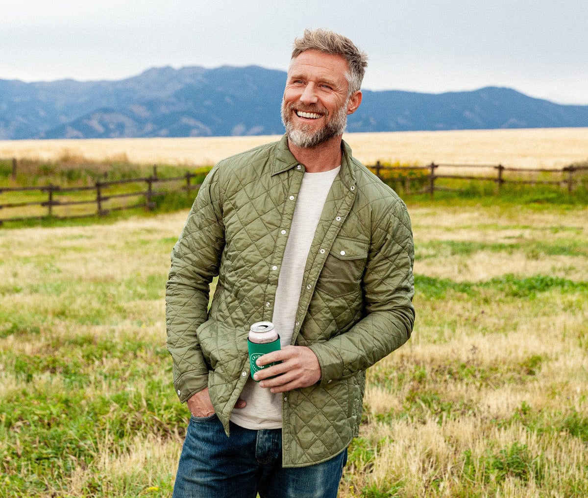 A smiling bearded man stands outdoors in a grassy field, wearing the CRIQUET Quilted Shacket by CRIQUET, paired with jeans and holding a canned drink. Mountains and a wooden fence can be seen in the background.
