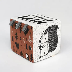 Introducing the WEE GALLERY - WOODLAND SOFT BLOCK by WEE GALLERY, a soft fabric block adorned with a whimsical design perfect for stimulating a baby's developing senses. One side features a black and white squirrel holding a nut, while other sides showcase trees, stars, and owl illustrations in black, white, and brown. Small grey fabric tags protrude from the top for added sensory exploration.