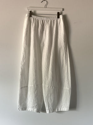 CUT LOOSE - LANTERN PANT by CUT LOOSE are white, wide-leg linen pants with an elastic waistband, hanging from a wooden hanger on a neutral-colored wall. These lightweight, breathable pants have a relaxed fit and feature visible seams at the knees.