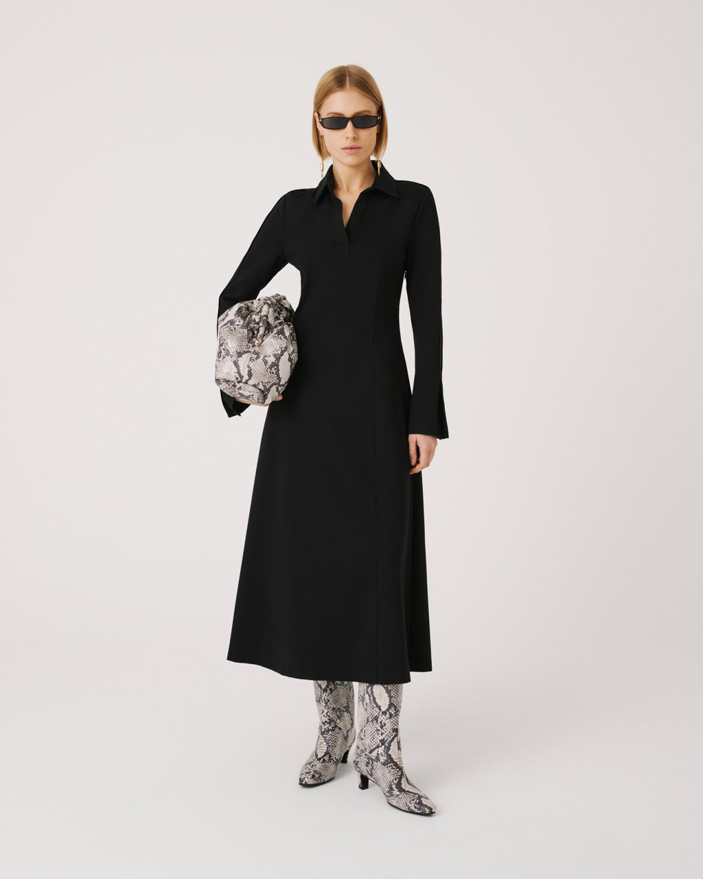 A person with short blond hair stands facing away, wearing a long black coat and snakeskin textured high-heeled boots. The individual, clad in dark sunglasses and the SFIZIO Ponte Knit Midi Dress with long sleeves and a split neck collar, is set against a plain light gray background.