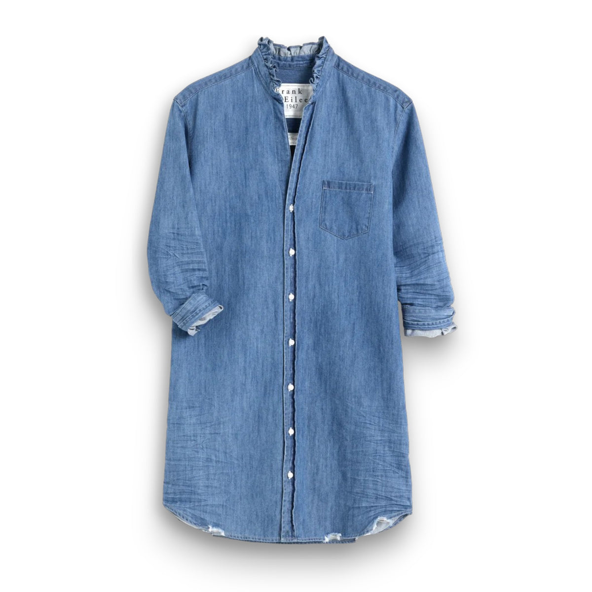 A woman poses, smiling on a stone patio with curly hair flowing freely. She wears the FRANK &amp; EILEEN Mary Ruffle Shirtdress in Vintage Stone Washed Indigo, set against ornate doors and a wall-mounted lamp offering an elegant backdrop.