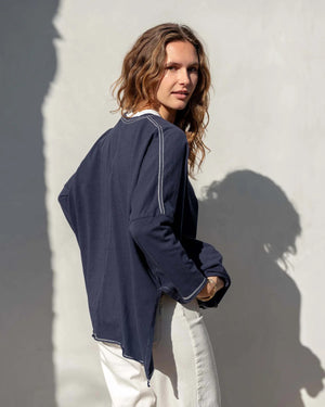 A person stands against a plain white wall, looking at the camera. They have curly hair pulled back and are wearing a loose-fitting, dark gray MERSEA Catalina Slub Long Sleeve Tee by MER SEA with 3/4 length sleeves, blue jeans, and a gold necklace with a circular pendant. Their shadow is visible on the wall behind them.