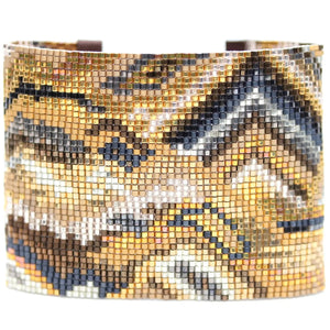 Introducing the JULIE ROFMAN - ORO CUFF by JULIE ROFMAN JEWELRY, a beaded bracelet that showcases an intricate geometric pattern in hues of gold, brown, blue, and white. The delica glass beads form a mosaic-like effect with abstract mountain and wave motifs in a stunning landscape design accented with 24kt gold-plated touches.