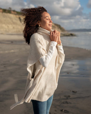 A person standing against a plain wall is dressed in the MERSEA Cape Poncho Sweater, made from a beige cotton knit blend, layered over a brown long-sleeve shirt and paired with dark blue pants. With one hand resting over the other, the poncho’s loose fit and ribbed hem provide both comfort and style.