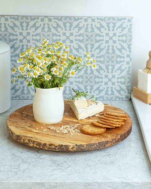 A small white vase filled with yellow and white daisy flowers sits on a PETERMANS BOARDS & BOWLS INC SPENCER PETERMAN - SPALTED LAZY SUSAN 20" along with a wedge of soft cheese topped with herbs and a few round crackers. The spinning tray rests on a kitchen counter, showcasing a patterned tile backsplash in the background.