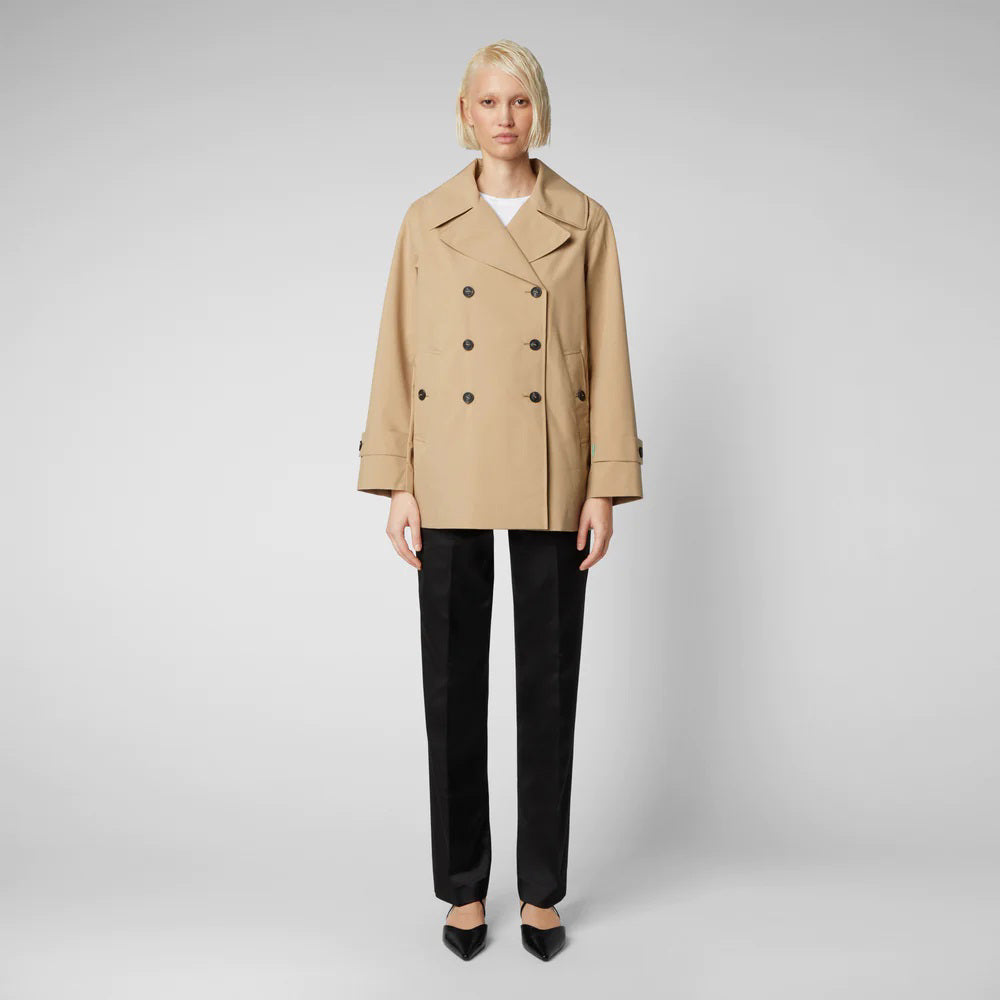 An individual with short blonde hair is wearing the SAVE THE DUCK - SOFI TRENCH JACKET, a beige, weather-resistant double-breasted coat. They are also dressed in a white top, black pants, and black pointed shoes. They are standing against a plain gray background, facing forward with arms at their sides.