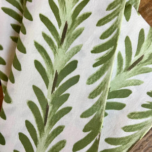 The CHRISTINA DICKSON HOME FERN COTTON NAPKIN, featuring a green leaf block print on 100% cotton and held by a brown ring, complements the matching tablecloth beneath with its seamless leaf motif design. A small green plant is visible in the top left corner.