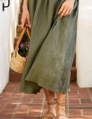 A person wearing the FRANK AND EILEEN - DAPHNE SLEEVELESS POPOVER DRESS in a flowing, olive-green hue stands on brick steps. They hold a small woven basket filled with purple flowers and a bit of greenery. Only their hand, part of their forearm, and their legs in strappy sandals are visible.