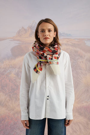A woman with light brown hair wears a cream shirt and the STORIATIPIC - ANOUK WOOL SCARF, an elegant hand-embroidered accessory by STORIATIPIC, wrapped around her neck. Mountains and plains form a soft, scenic backdrop.