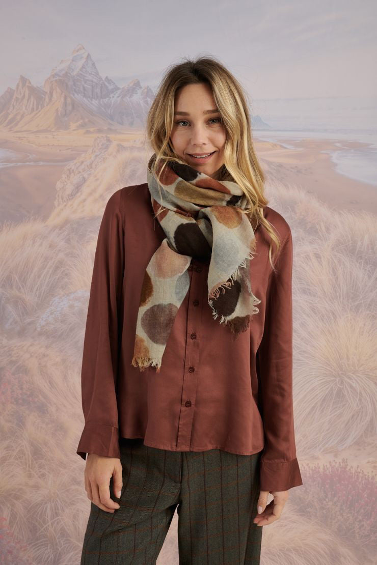A woman with blonde hair displays the STORIATIPIC - BETTY WOOL SCARF, a large rectangular wool accessory with earth-toned polka dots, set against a scenic mountain landscape.