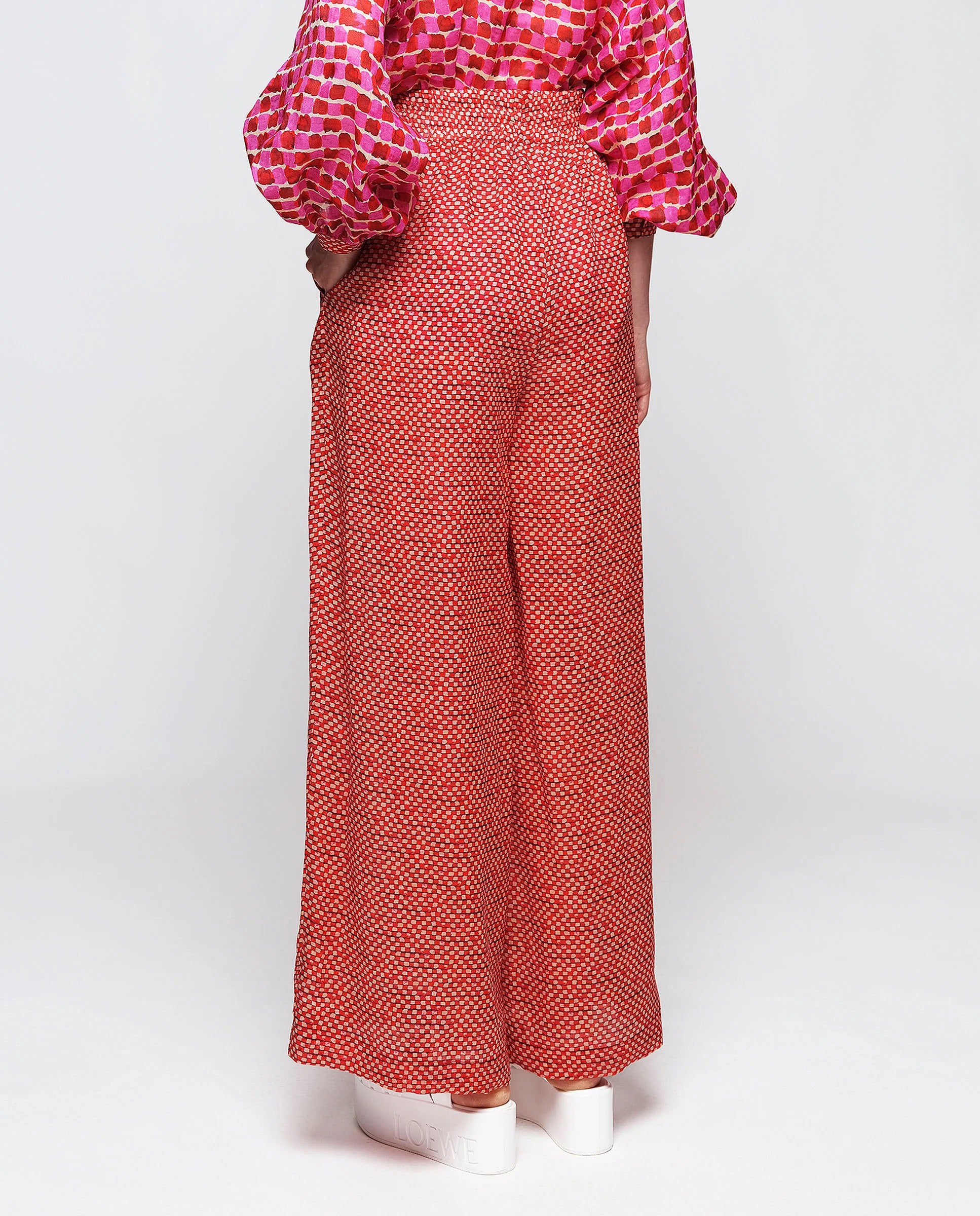 A person wearing the MIRTO 1956 - MOSAIC RAMIE TROUSER, featuring a high-waisted, wide-leg design with an elastic waistband and a small geometric pattern. The trousers are paired with a matching patterned top with billowy sleeves from MIRTO 1956. Completing the stylish yet comfortable look, the person is also wearing white platform sneakers.