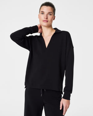 A person with dark hair styled in a neat updo is wearing the SPANX - AirEssentials Split Neck Polo Top in black, paired with matching black pants. They rest one hand on the back of their neck and gaze to the side against a plain white background, exuding an air of luxurious comfort.